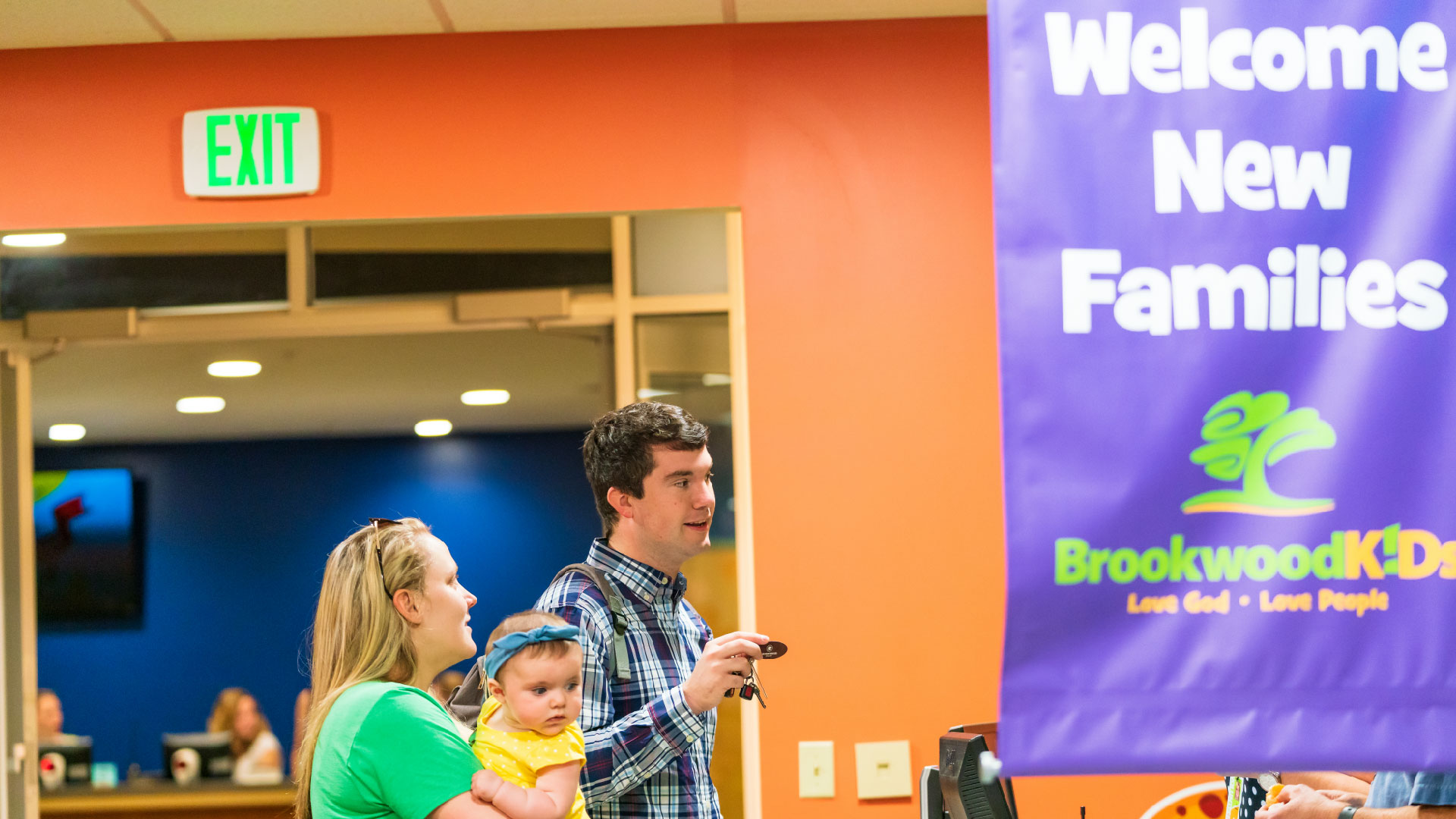 A family with a baby stands in front of the Welcome New Families banner at Brookwood Church